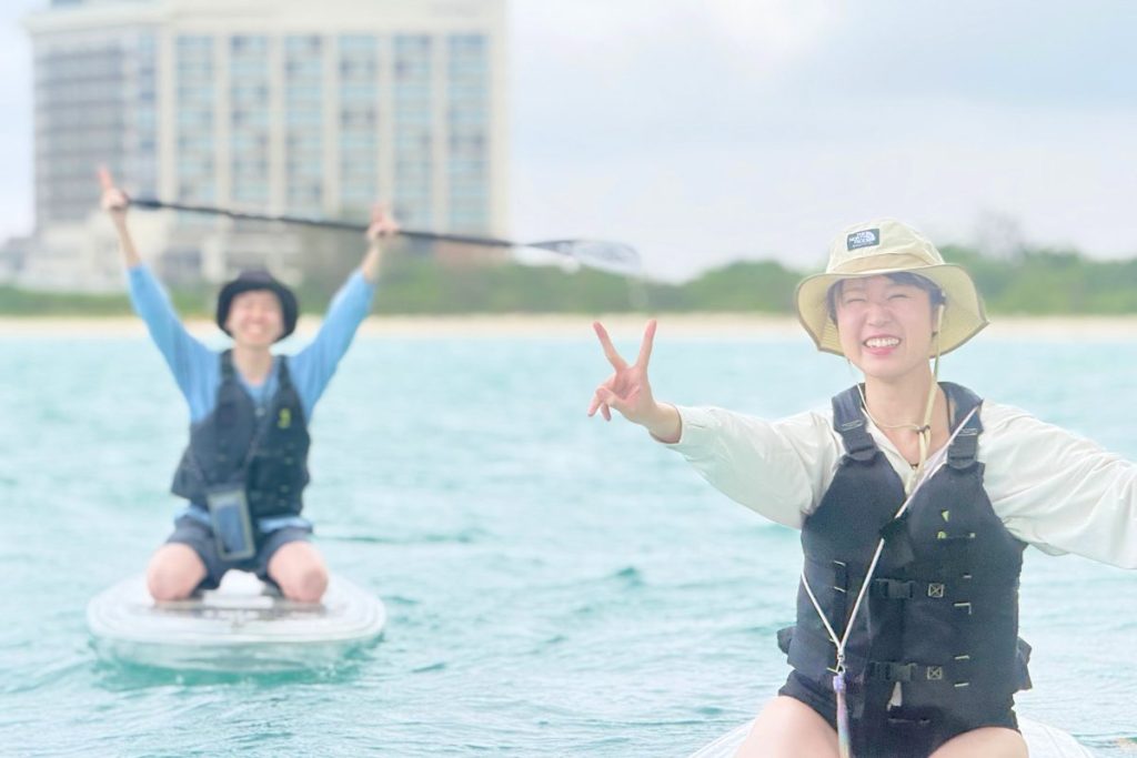 宮古島の海でクリアSUPを楽しむあやこさんとたくとさん。手前で笑顔でピースするあやこさんと、後ろでパドルを高く掲げるたくとさん