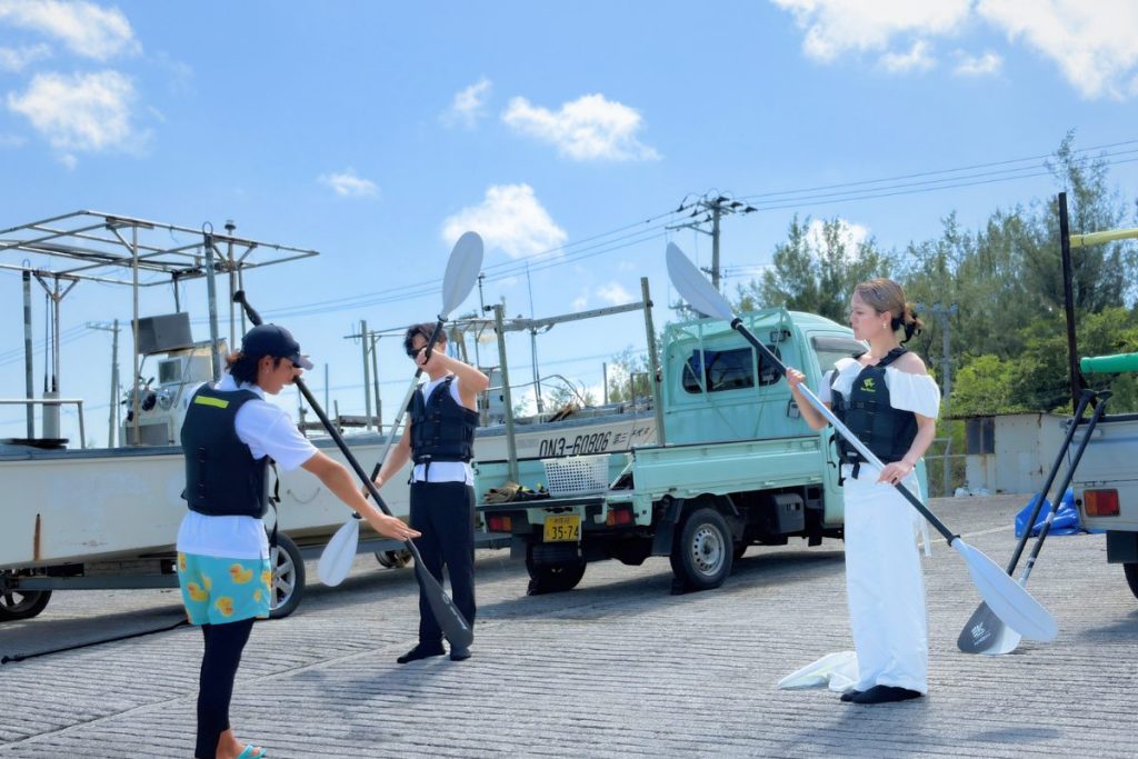 ユニの浜へ向かう前に、ウェディング衣装の新郎新婦がガイドのしょーだいからSUPパドルの使い方を教わる様子