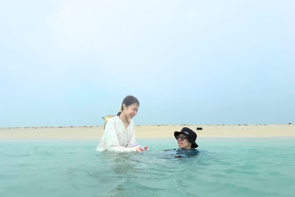 ユニの浜の浅いエメラルドグリーンの海に入り、砂浜を背景に笑顔で話すあやこさんとたくとさん