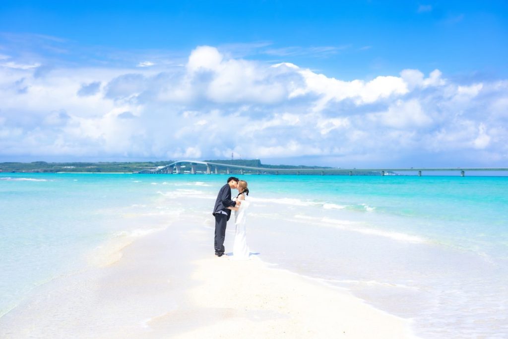 宮古島・ユニの浜の白い砂浜で、エメラルドブルーの海と伊良部大橋を背景にキスをする新郎新婦のフォトウェディング