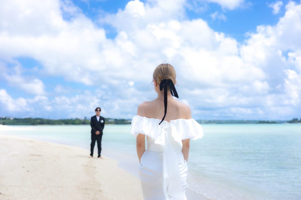 ユニの浜の白い砂浜で、ウェディングドレス姿の新婦が少し離れた場所に立つ新郎を見つめるロマンチックな前撮り写真