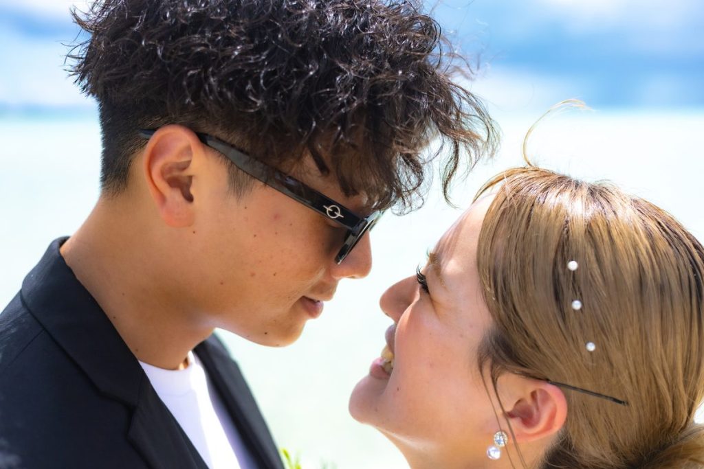 ユニの浜の青い海を背景に、黒いスーツ姿の新郎とウェディングドレス姿の新婦が見つめ合い笑顔を見せるアップのウェディングフォト
