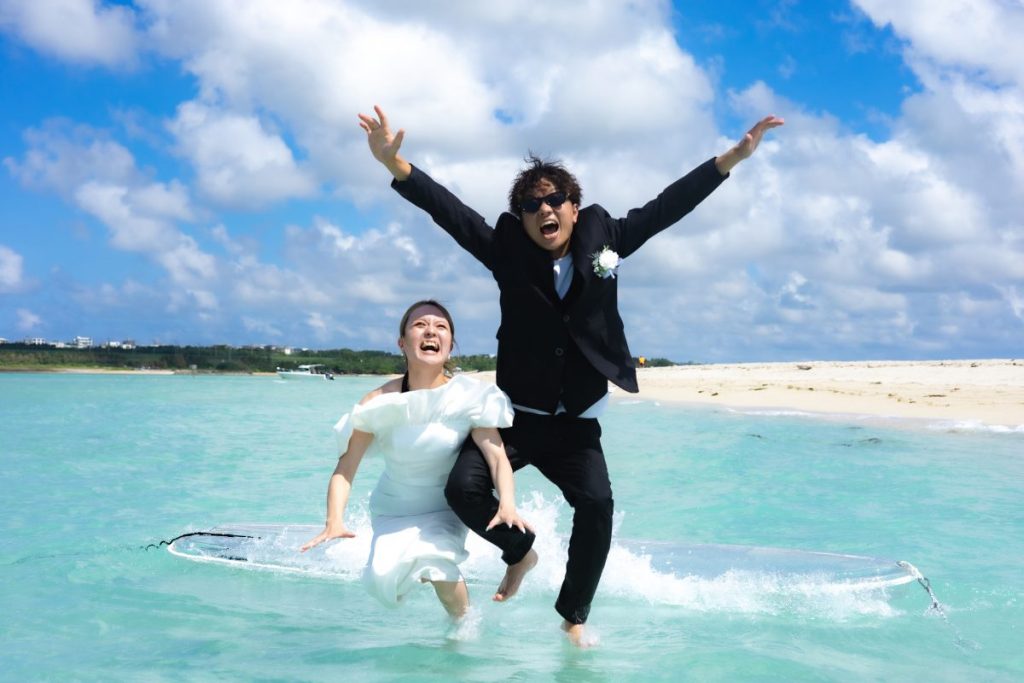 宮古島・ユニの浜の浅瀬で、ウェディング衣装の新郎新婦がクリアSUPの前から笑顔でジャンプする楽しげなフォトウェディング