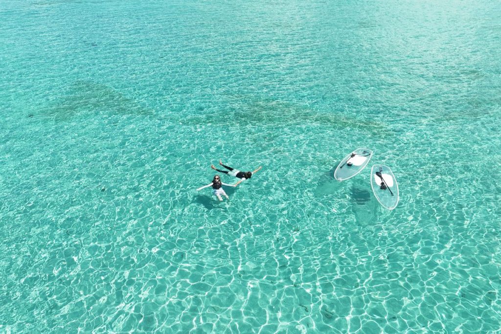 ユニの浜の透き通ったエメラルドブルーの海で泳ぐかすみさんとガイド、近くに2台のクリアSUPが浮かぶドローン写真