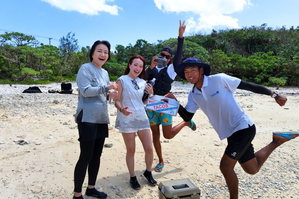 宮古島の秘境ビーチで、タコスの箱を囲んで笑顔で記念撮影するまあいさん・なつみさん親子とガイド二人